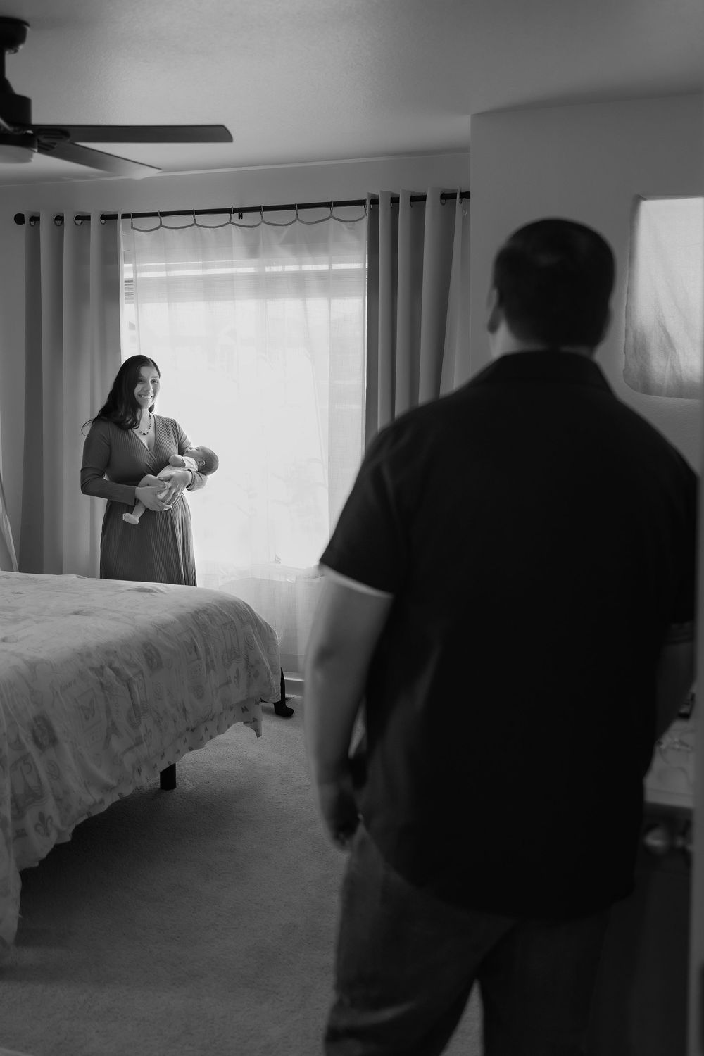 Black and white photo.  Mom stood next to window holding newborn baby, dad stood in the doorway looking in at mom, she is smiling at him