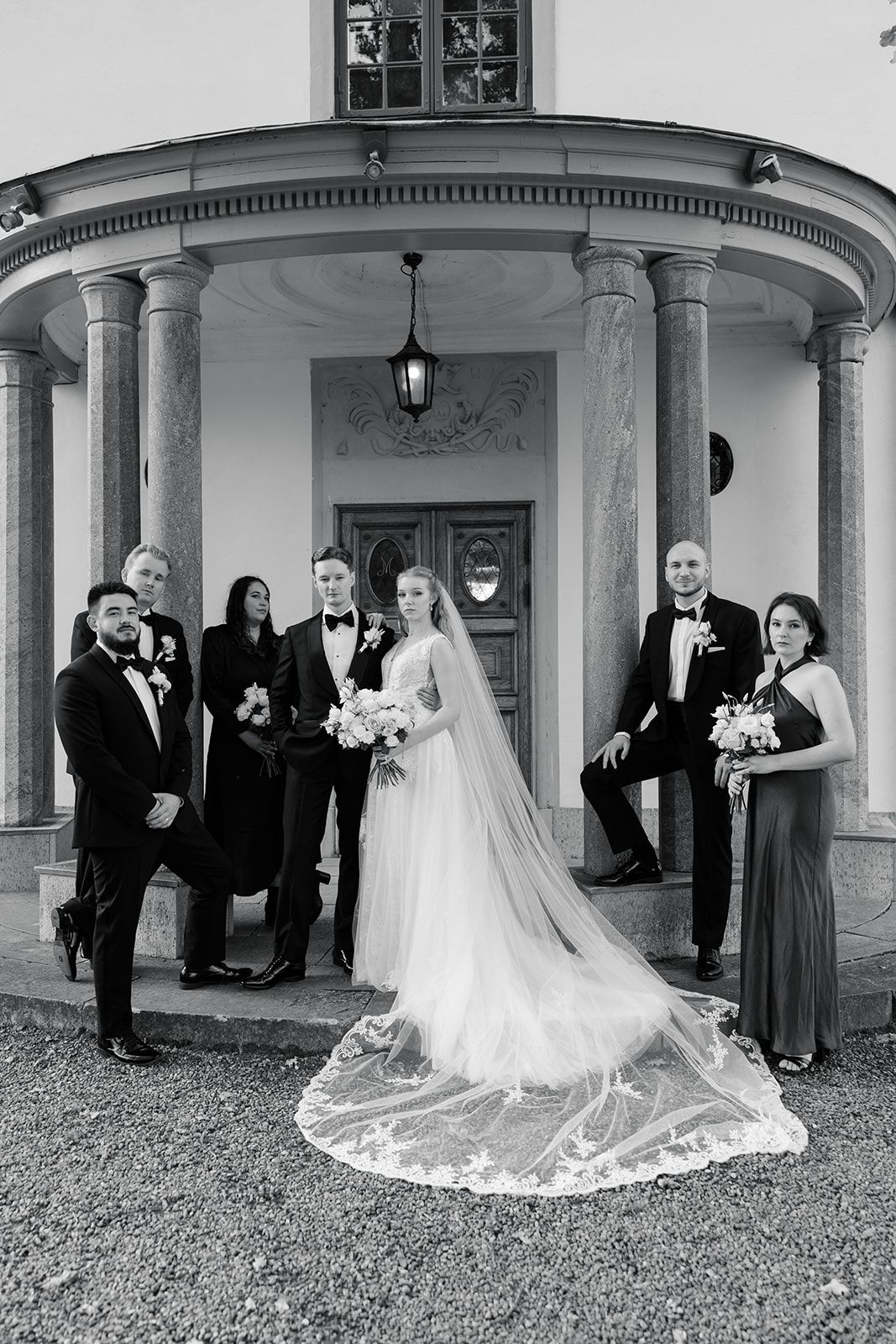 A wedding party gathers under an elegant columned portico with a sweeping white dress train on display.