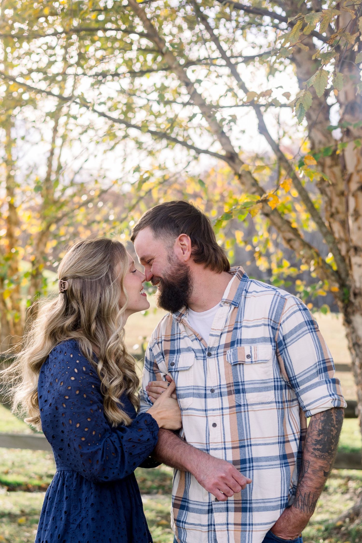 A couple in blue and plaid share tender moments outdoors during a fall photoshoot in a park setting.