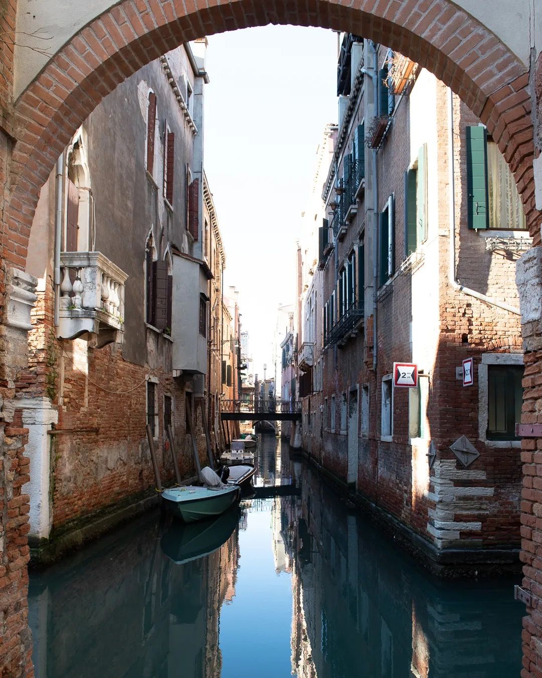 Venice canal framed by a brick archway with boats and historic buildings reflected in calm water.