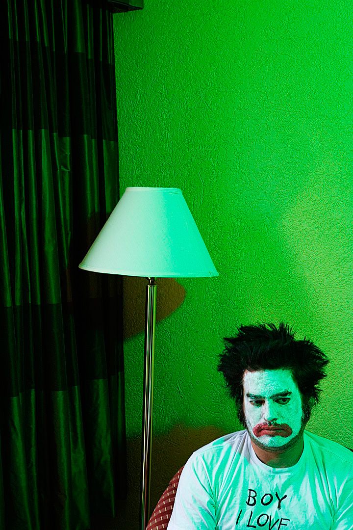 A person in a white t-shirt and face paint sits near a lamp with green lighting in a sequence of progressive shots.