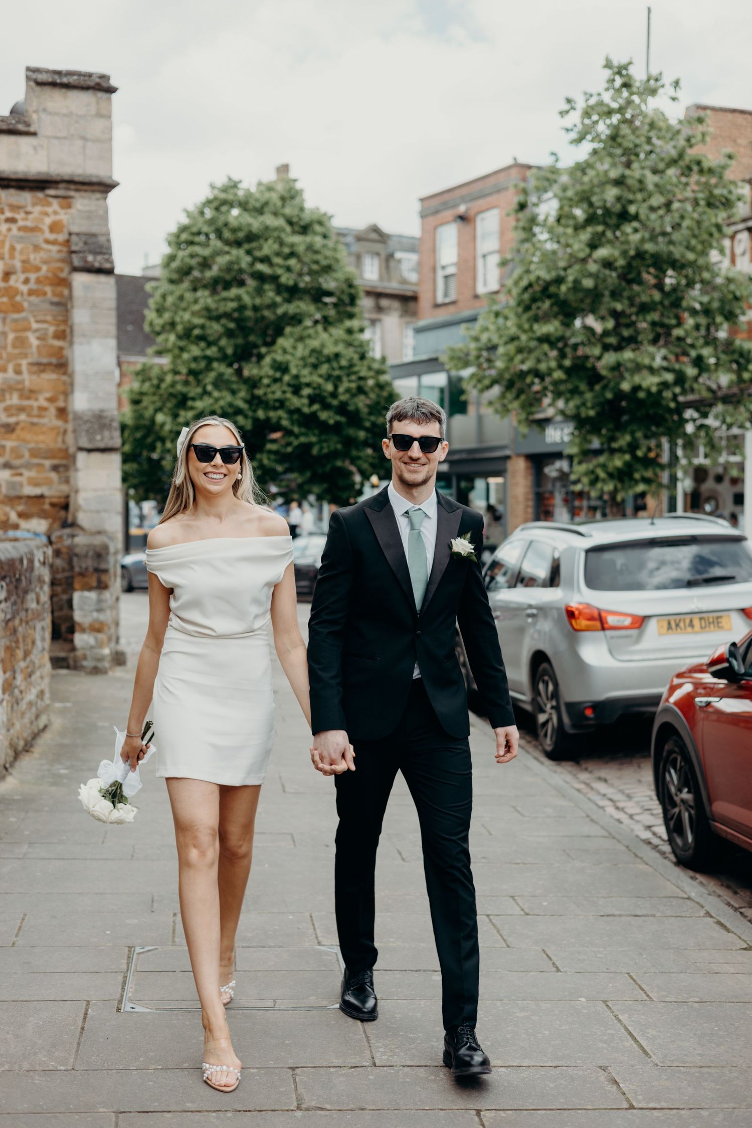 Stylish couple in wedding attire walks hand in hand down a city sidewalk with storefronts and parked cars in background.