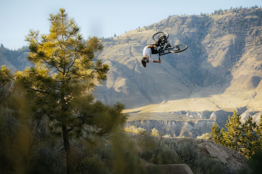 Mountain biker performs aerial jump trick against scenic mountain backdrop during sunset golden hour.