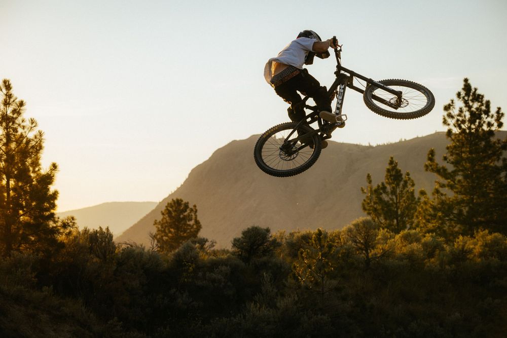 Mountain biker performs silhouetted backflip trick against sunset sky and mountain backdrop.