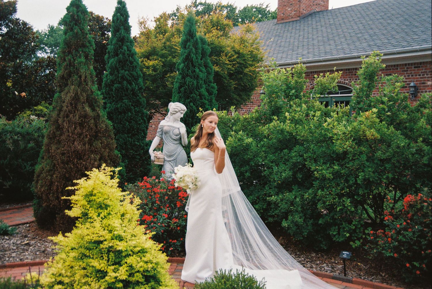 A wedding party walks along a brick pathway surrounded by lush green gardens and trees on a sunny day.