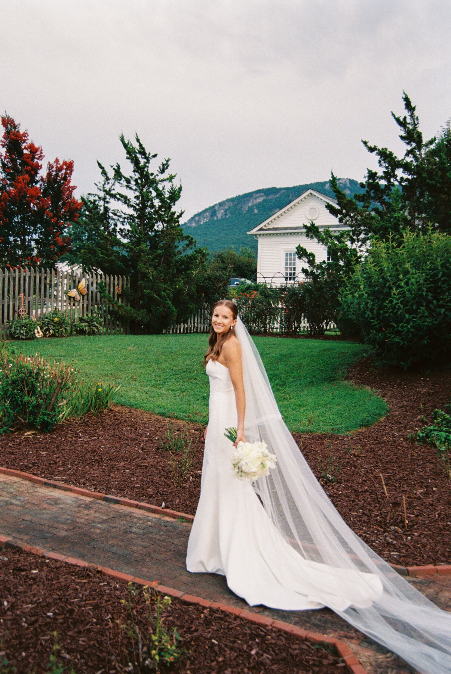 A bride in a flowing white gown and veil poses gracefully on a grassy hillside with mountain views and landscaped gardens.