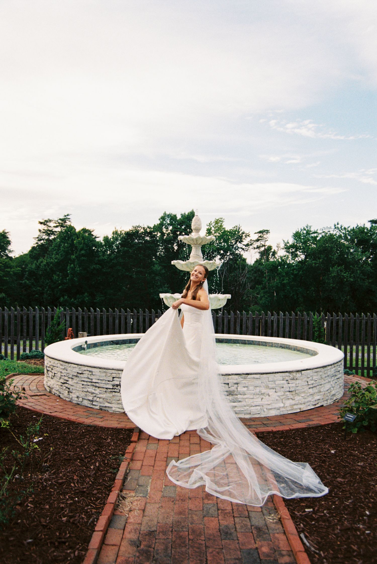 A bride twirls her flowing white dress and veil on a brick pathway surrounded by stone walls and landscaping.