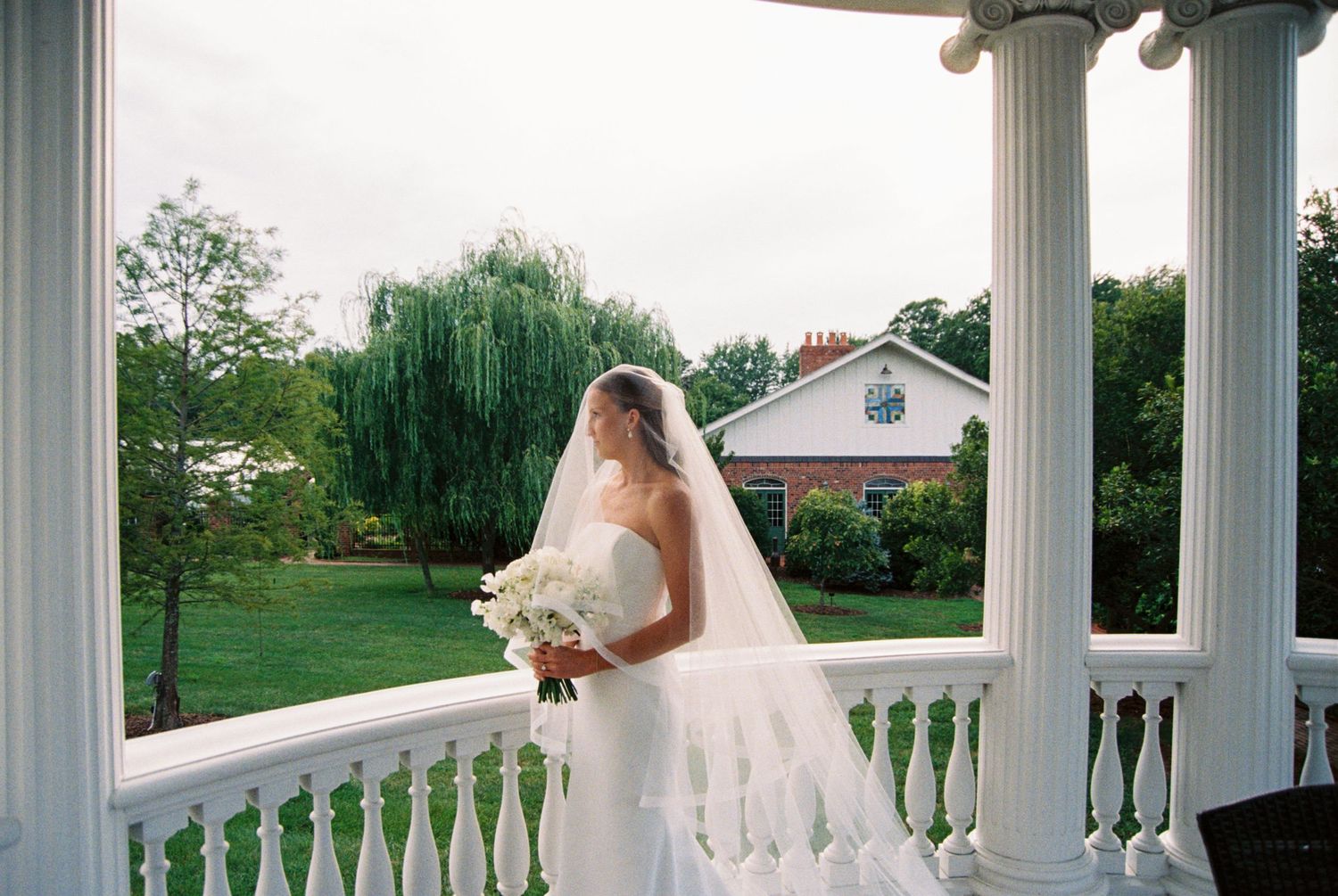 A bride in a white wedding dress and veil holds a bouquet while standing on a white columned porch with a garden view.