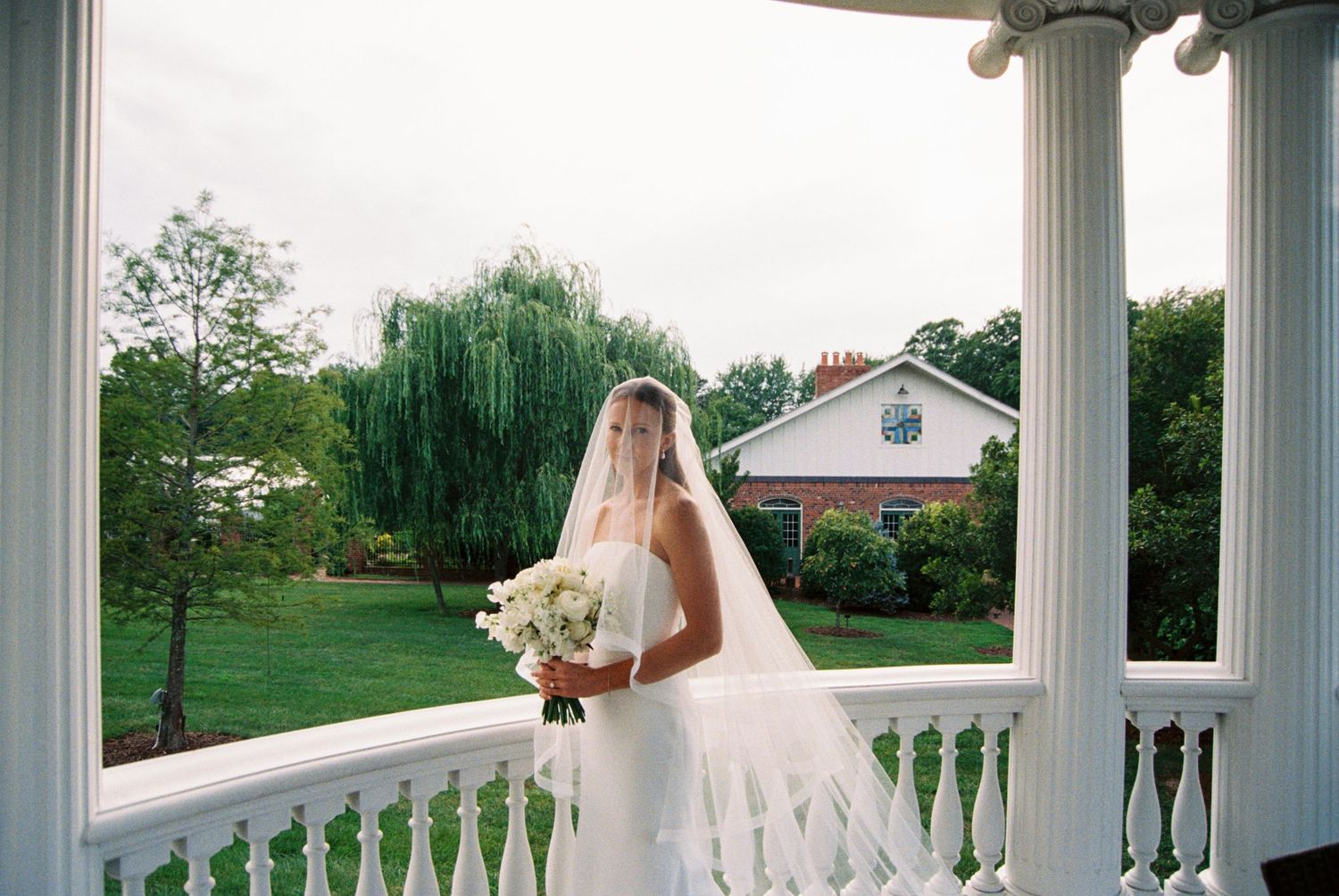 A bride in a flowing white dress stands between elegant white columns on a wraparound porch.