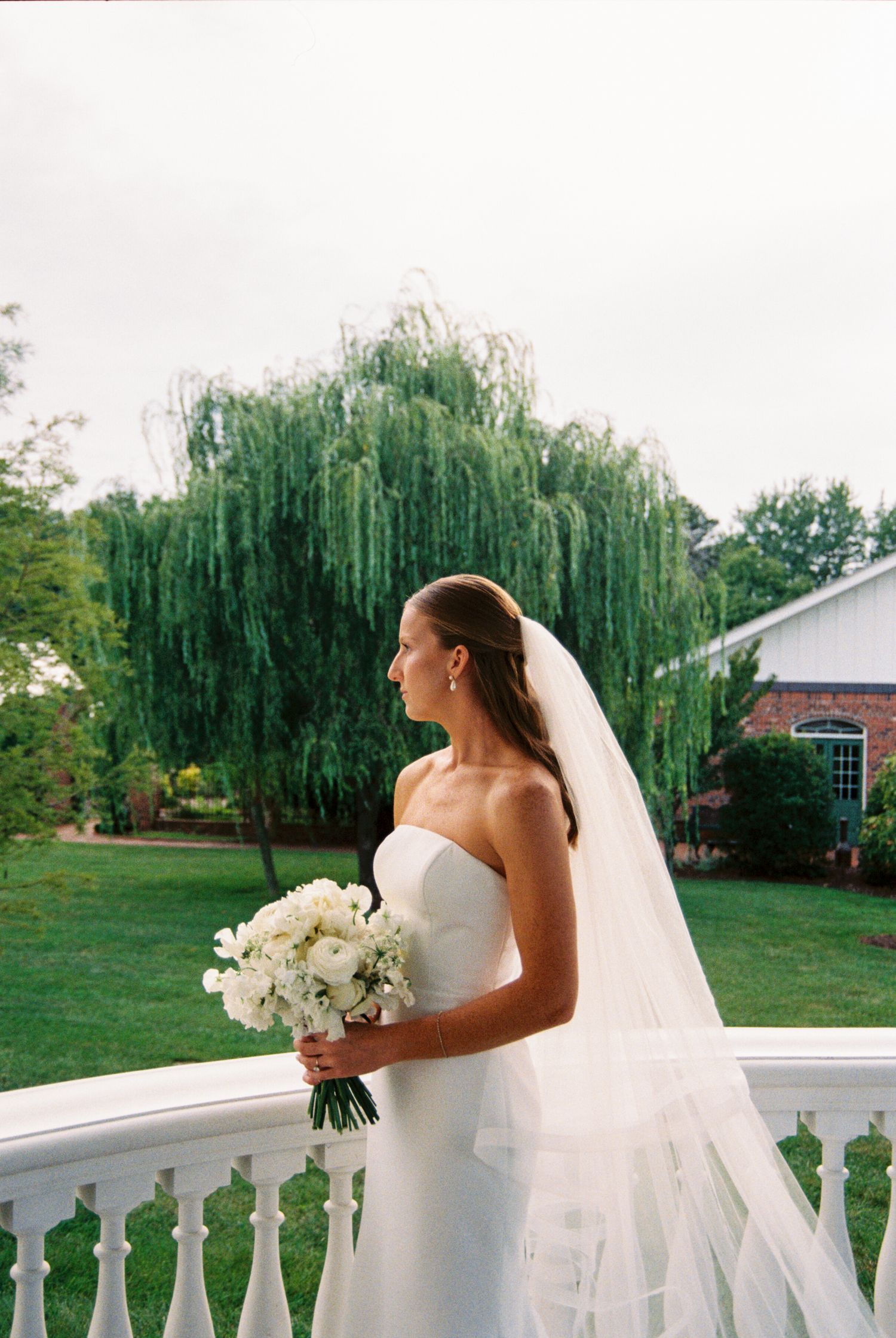 A bride in a strapless white gown stands with a bouquet on a porch with white railings and weeping willows in background.