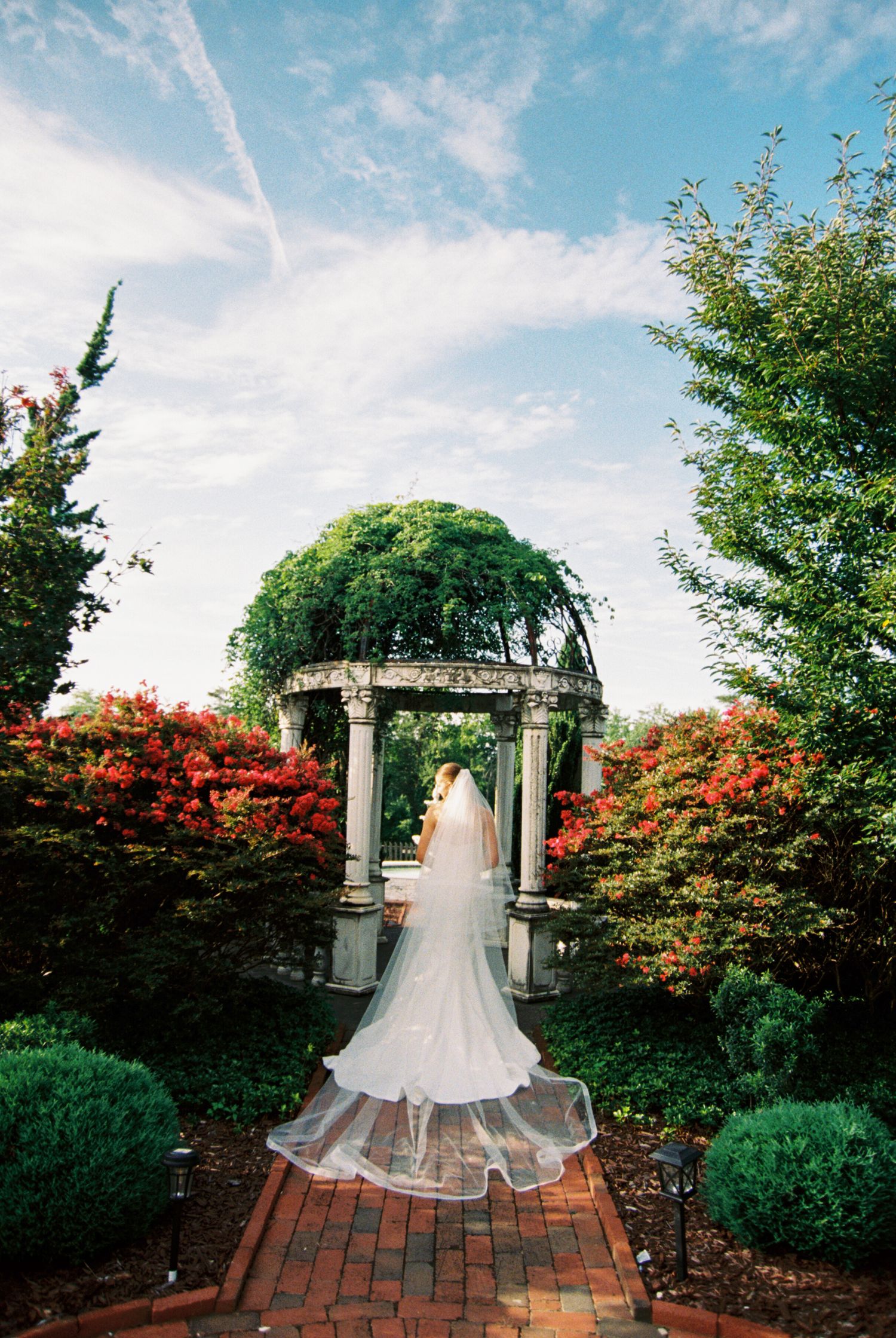 A wedding dress train flows elegantly across a brick pathway in a garden setting with stone pillars and red foliage.