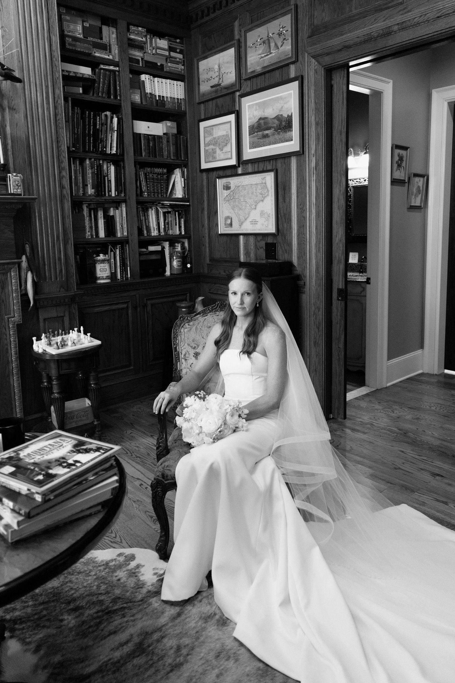 A bride in a white wedding gown sits in a cozy library setting with bookshelves and framed artwork on the walls.