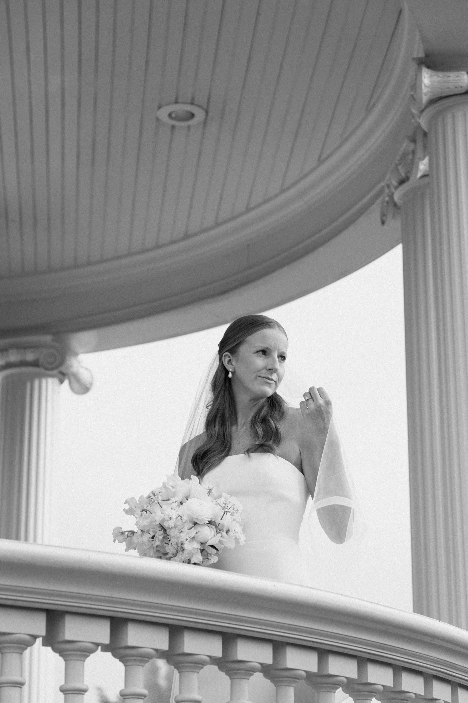 Elegant bride in white dress poses on a curved white balcony with floral decorations and classical columns.