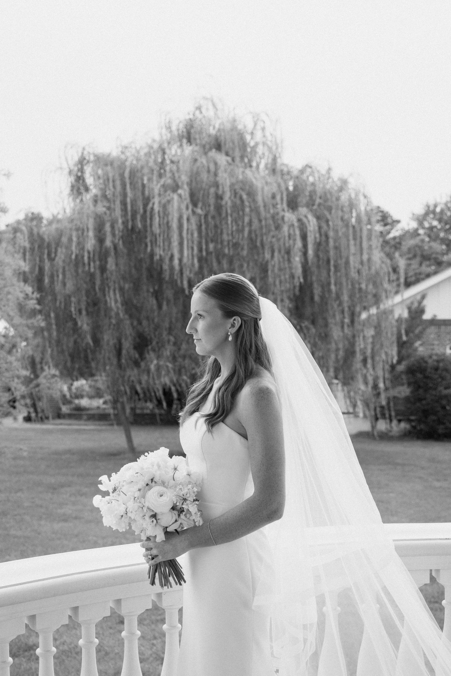 A bride in a strapless white gown stands on a white deck with willow trees in the background.
