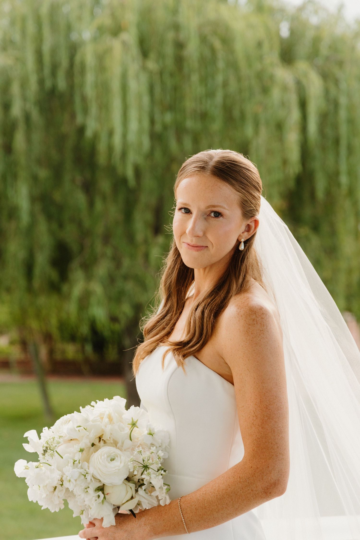Bride in white wedding dress holds elegant white orchid bouquet against backdrop of weeping willow trees.