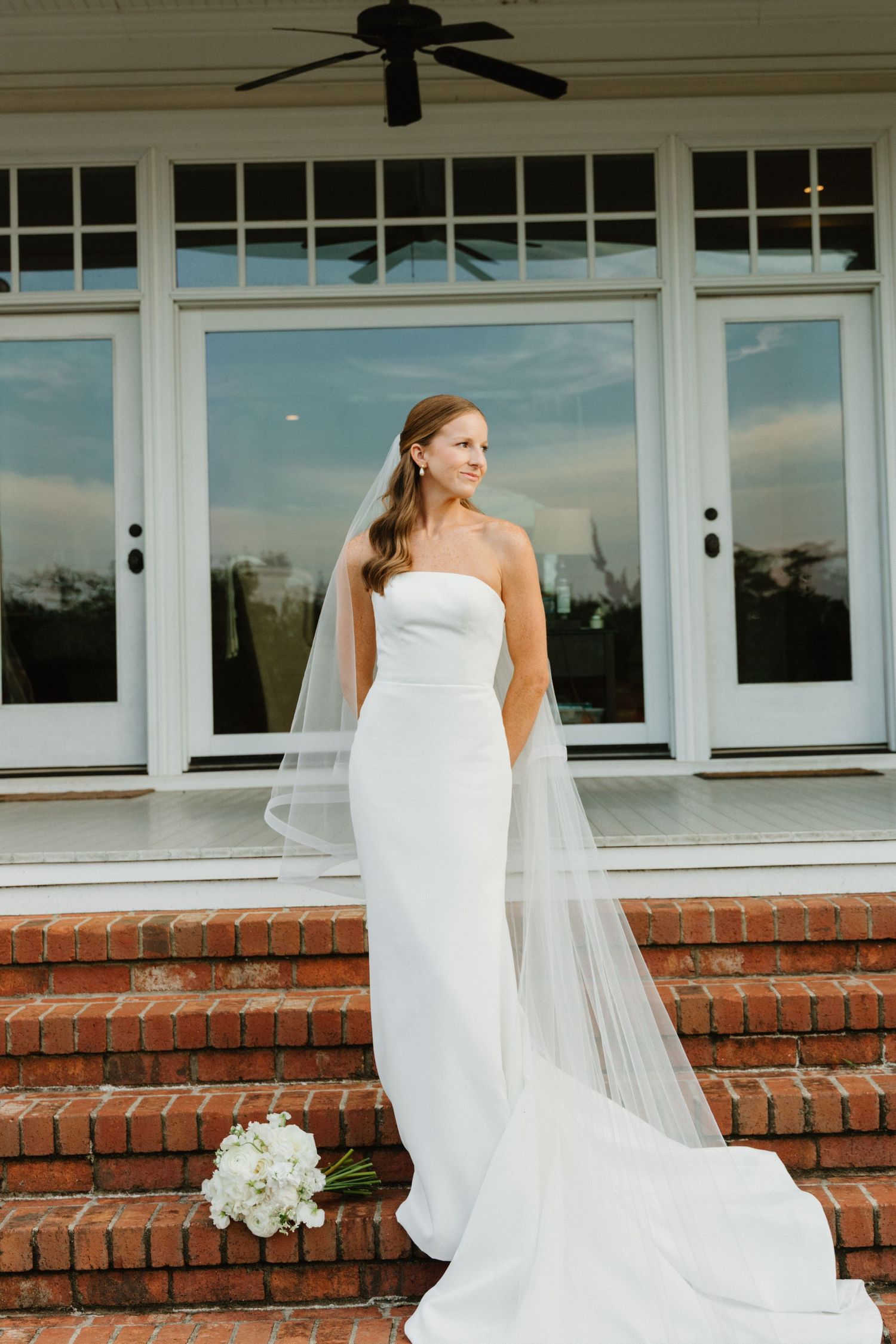 A bride in a white wedding dress and veil sits on a brick ledge outside a window-lined building.