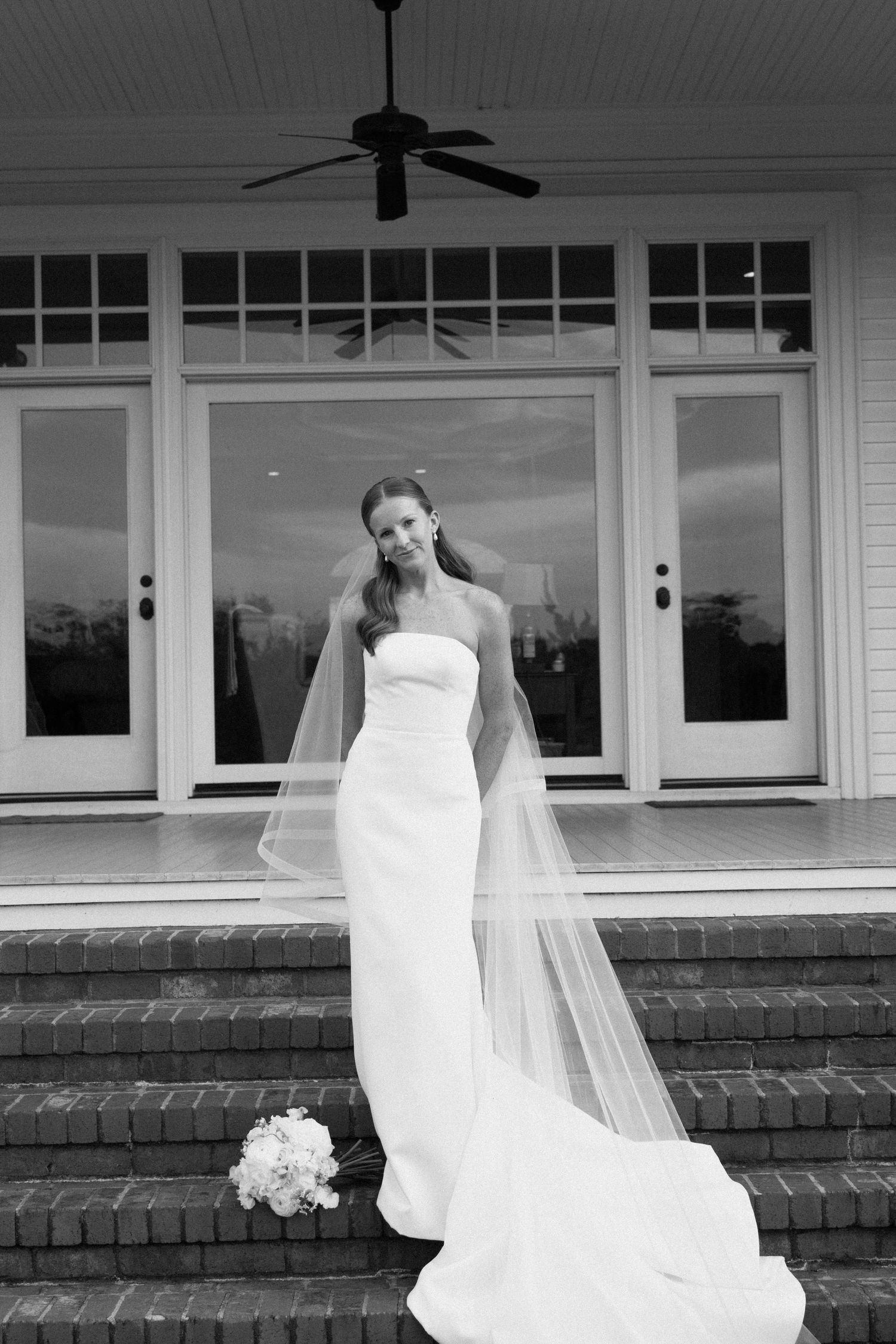 Black and white panoramic photo of a bride in a flowing white wedding dress and veil on a front porch.