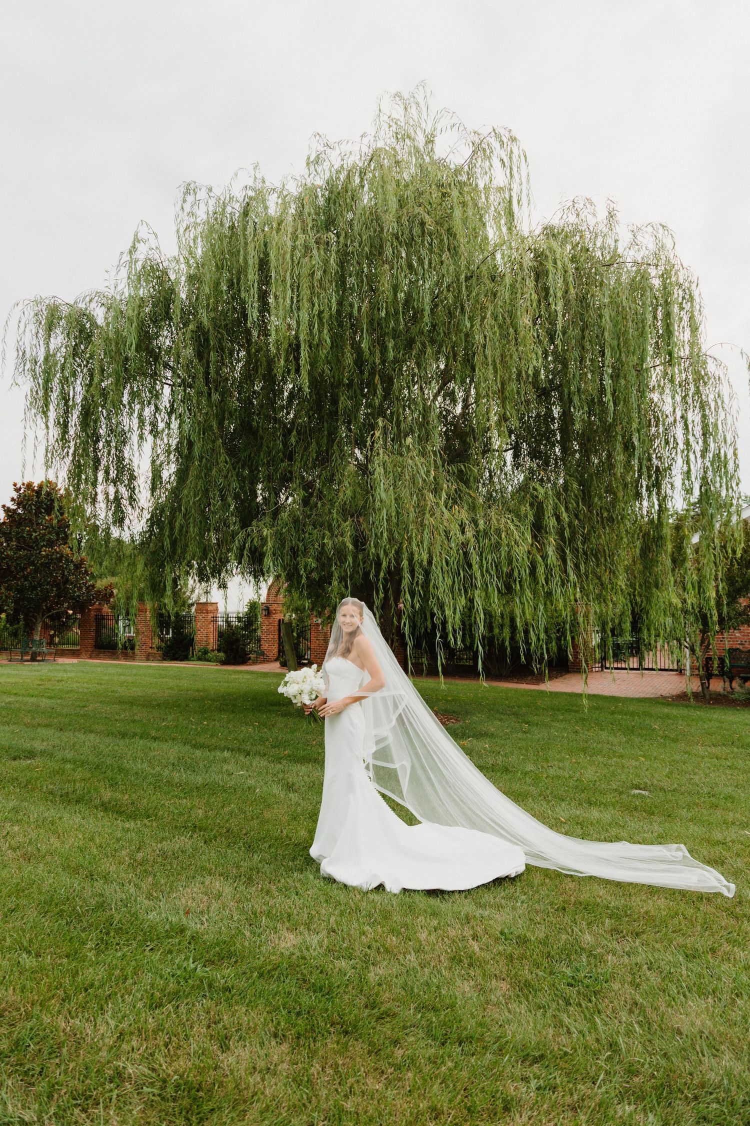 A bride in a flowing white wedding dress poses on a green lawn with cascading willow trees in the background.