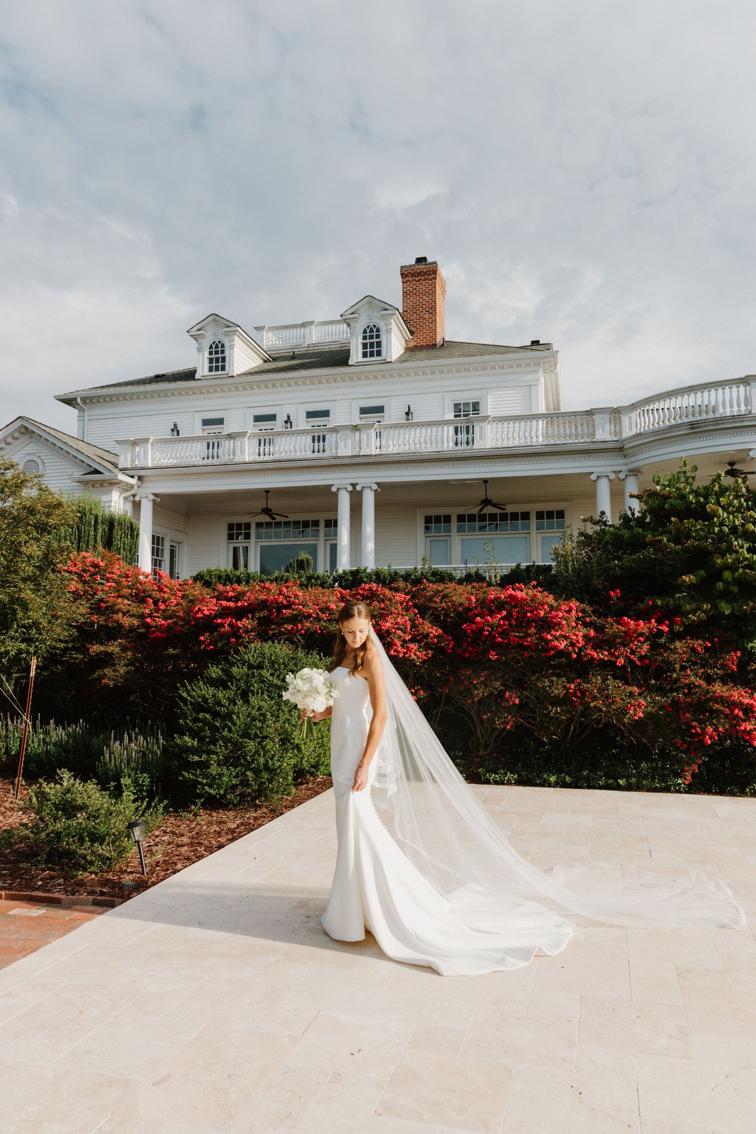 A bride in a flowing white wedding gown with a long cathedral train poses on a white terrace with red flowering bushes.