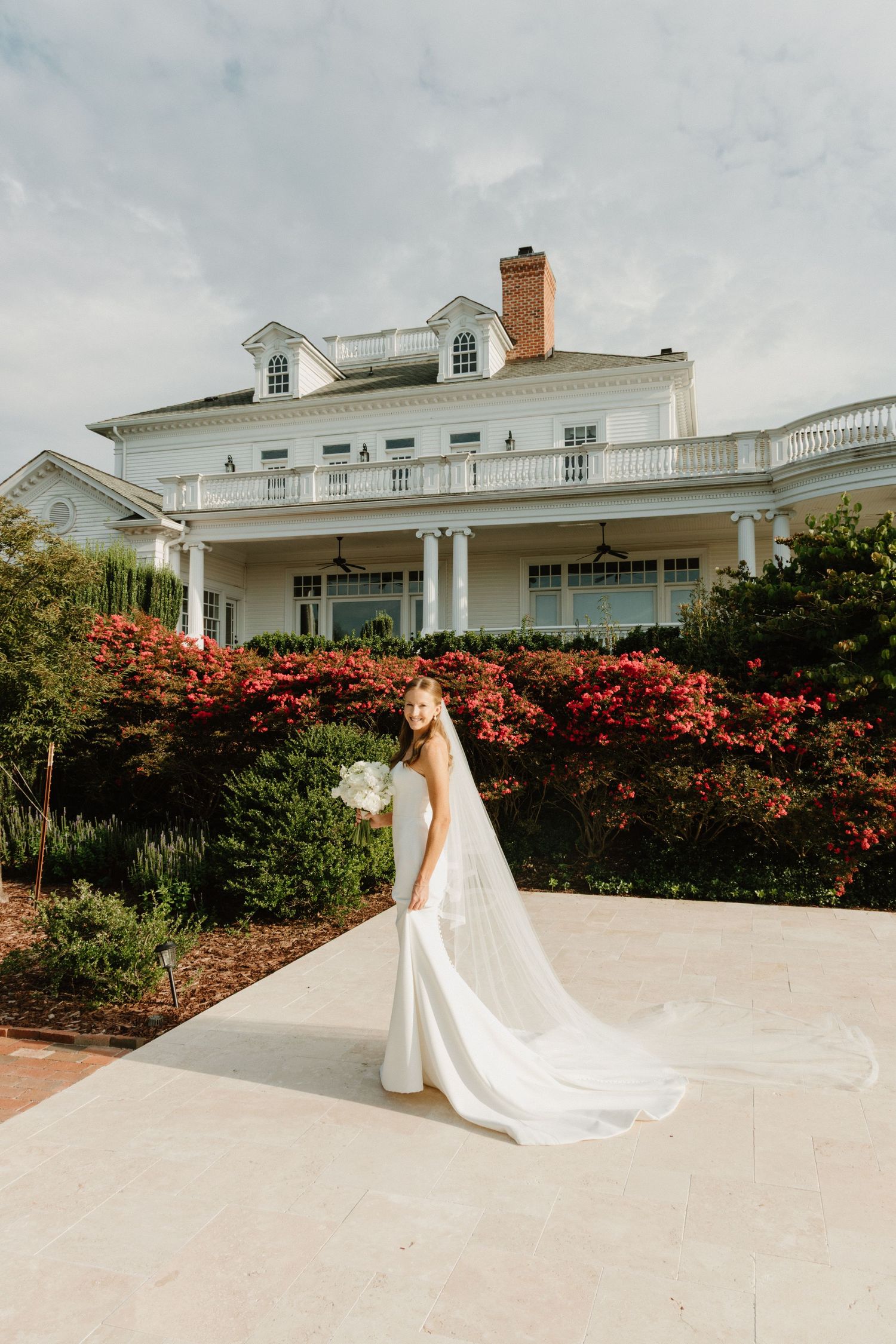 A person in a flowing white wedding gown poses in front of a stately white mansion with blooming red azalea bushes.