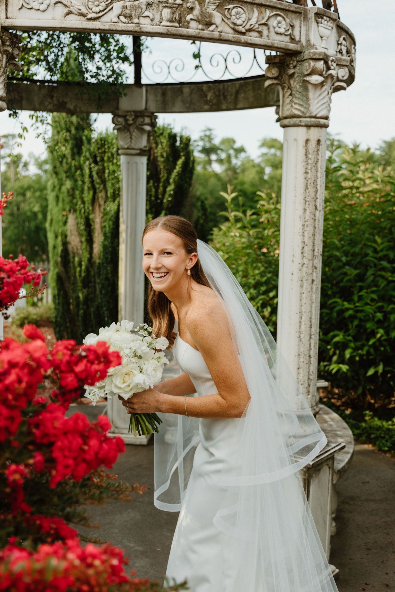 A bride in a white wedding dress smiles joyfully under an ornate stone gazebo surrounded by vibrant red flowers.