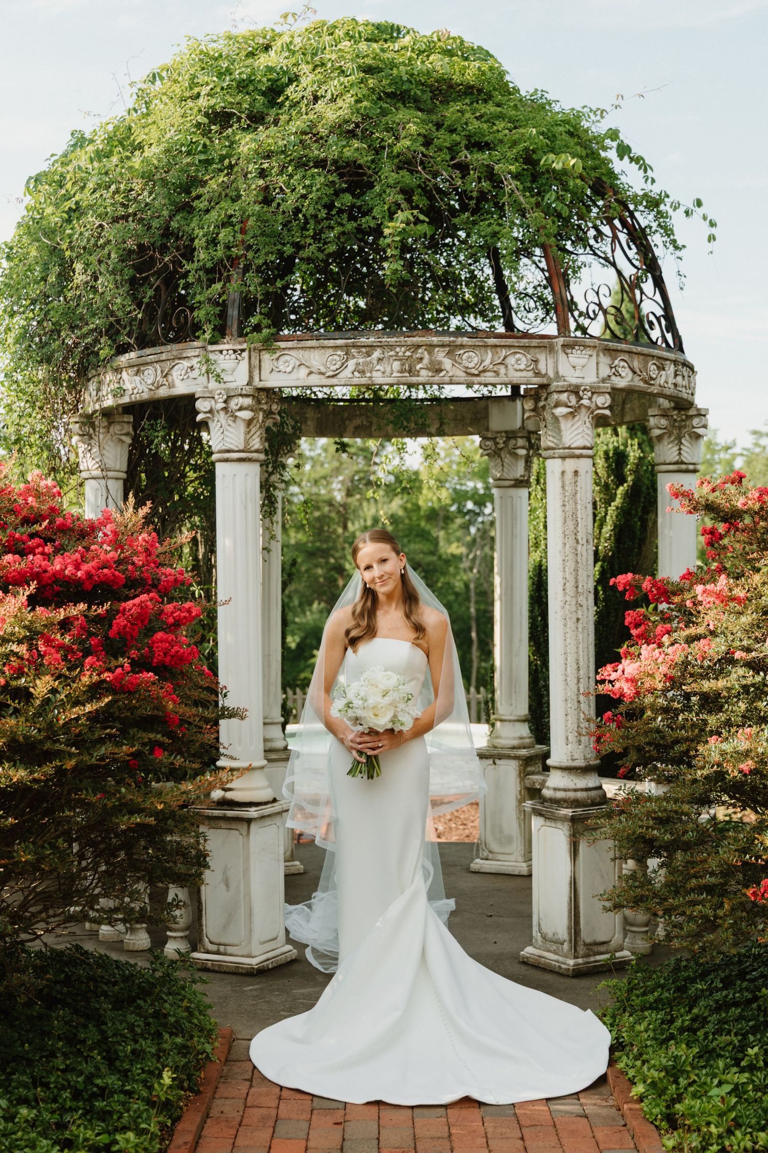 Elegant wedding photos captured in a garden setting with stone columns, lush greenery, and vibrant red azalea blooms.