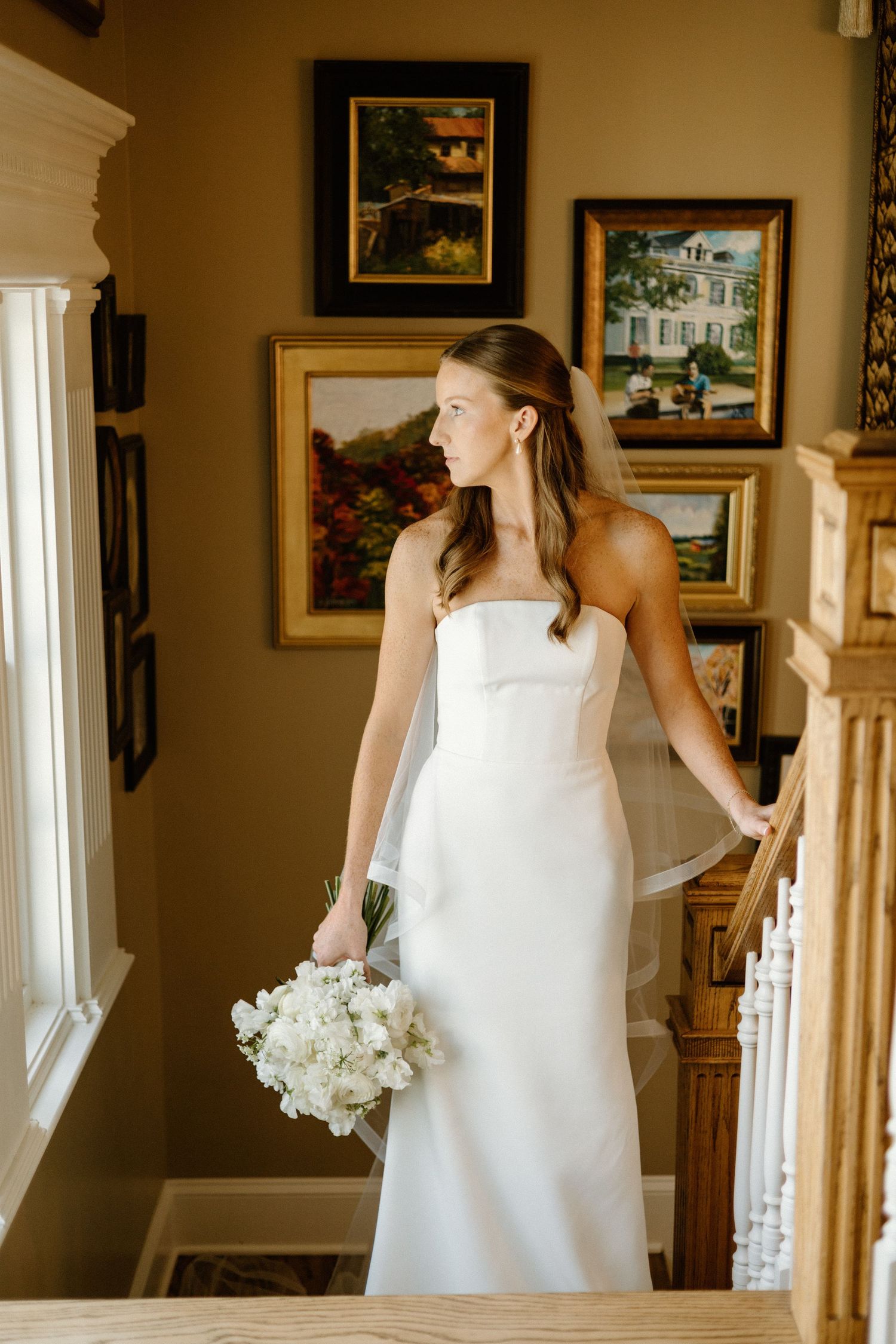 Elegant bride in white dress holding bouquet walks through hallway with framed artwork and white banister.