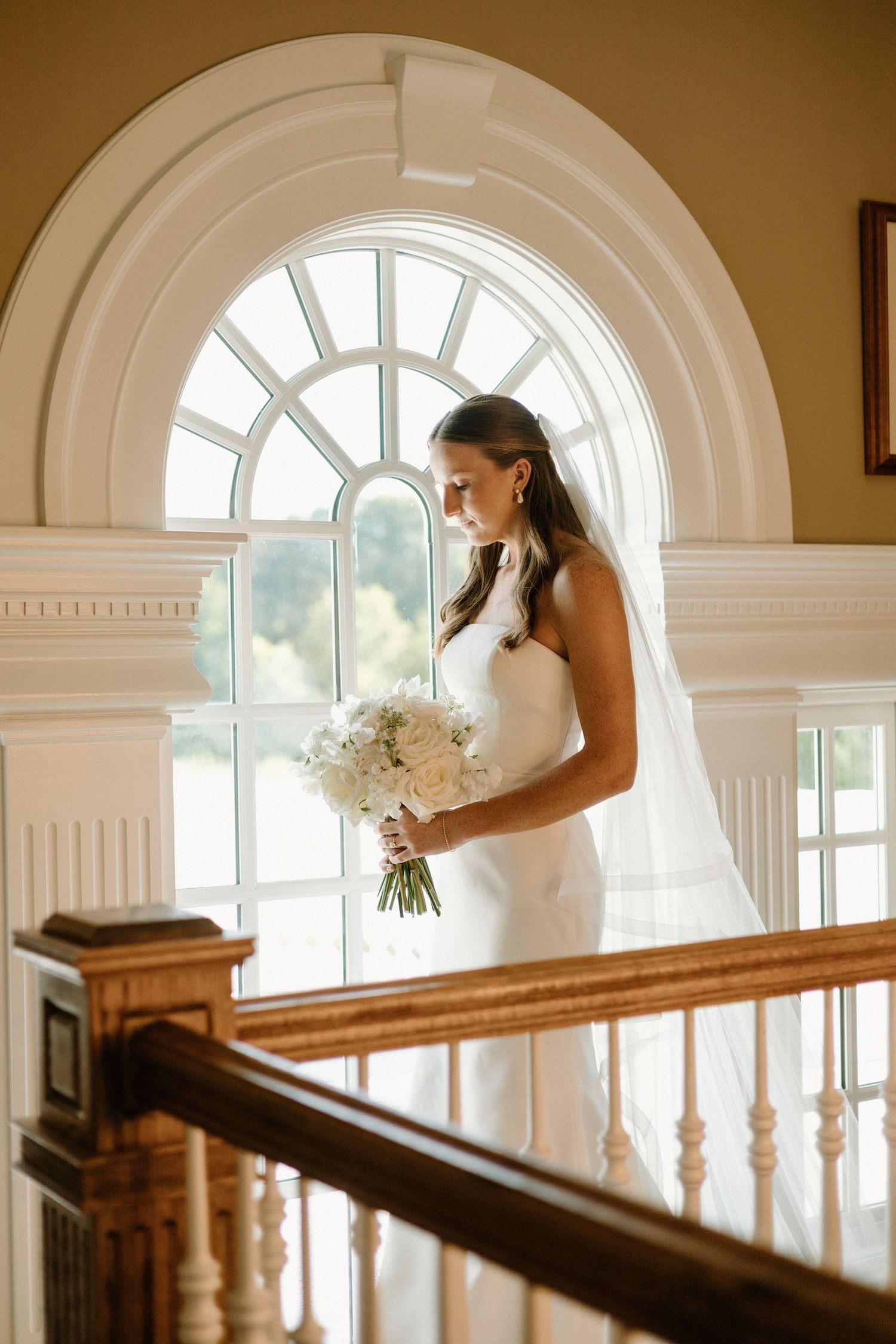 A bride in a white wedding dress stands by an elegant arched window holding a bouquet of white flowers.