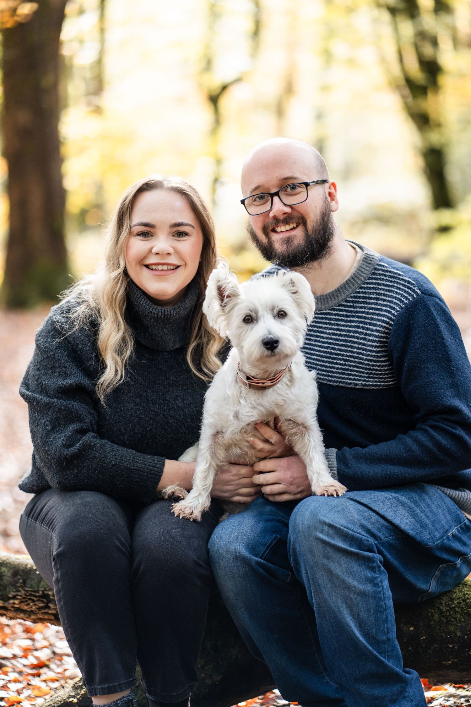 A couple sits outdoors in autumn with their small white dog amid fallen leaves and trees.