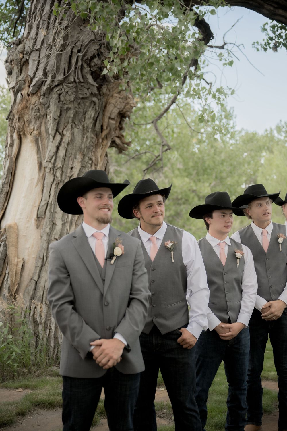 A group of groomsmen in matching black cowboy hats, gray vests, and dark pants stand together outdoors near trees.