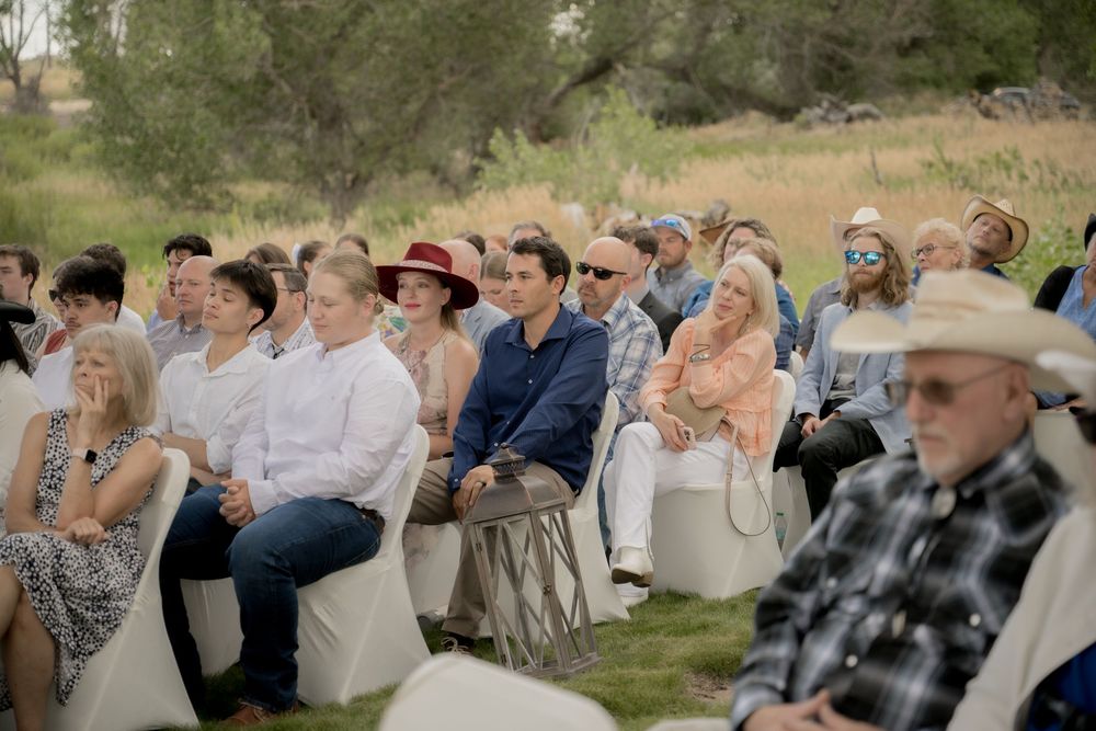 Wedding guests seated outdoors at a rustic ceremony watch attentively in a natural woodland setting.