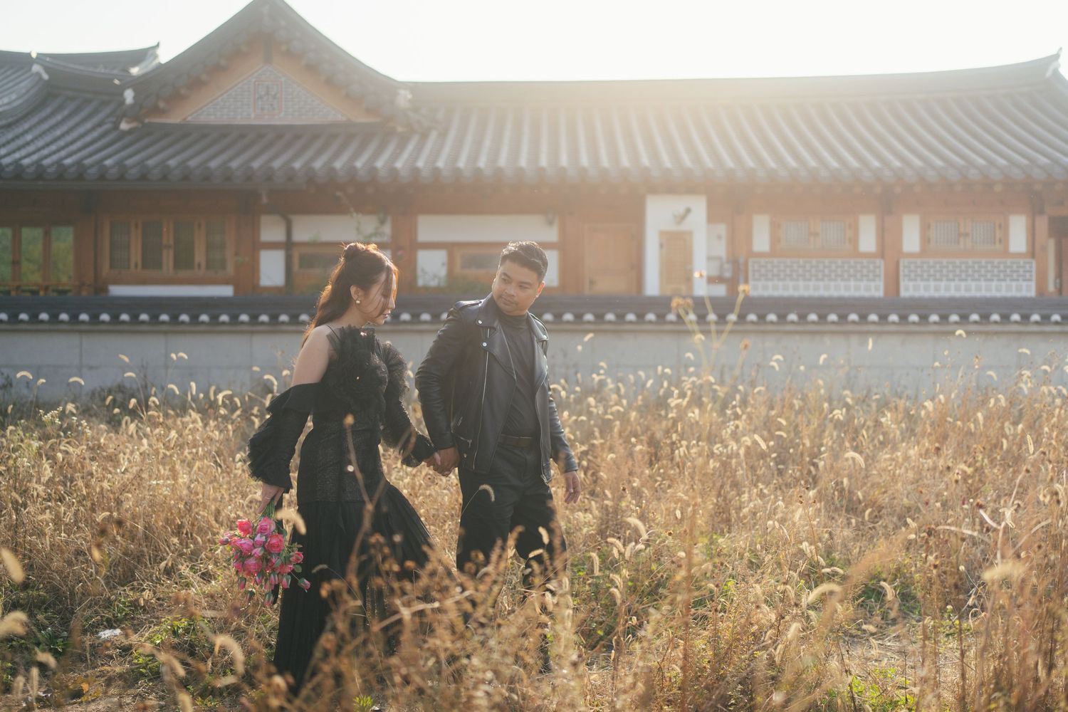 A couple walks through a golden field of tall grass near traditional Korean architecture at sunset.