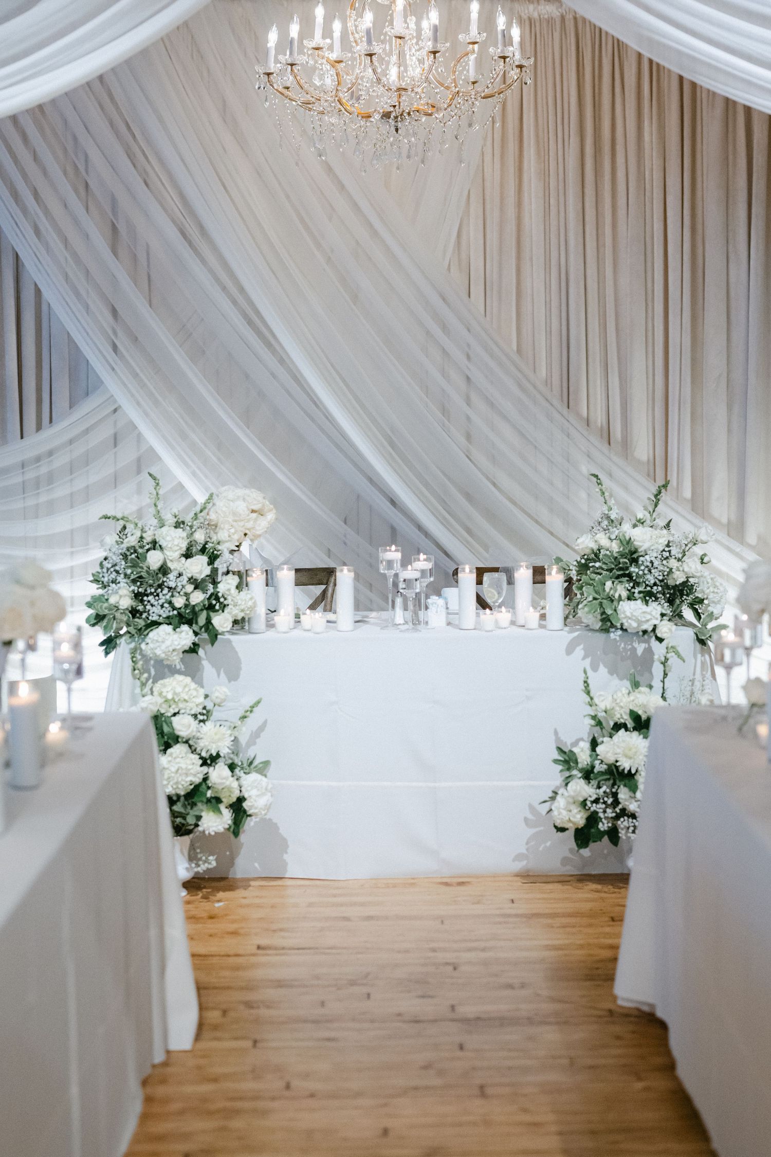 Elegant wedding reception venue with white draped ceiling, crystal chandeliers, and round tables on hardwood floors.