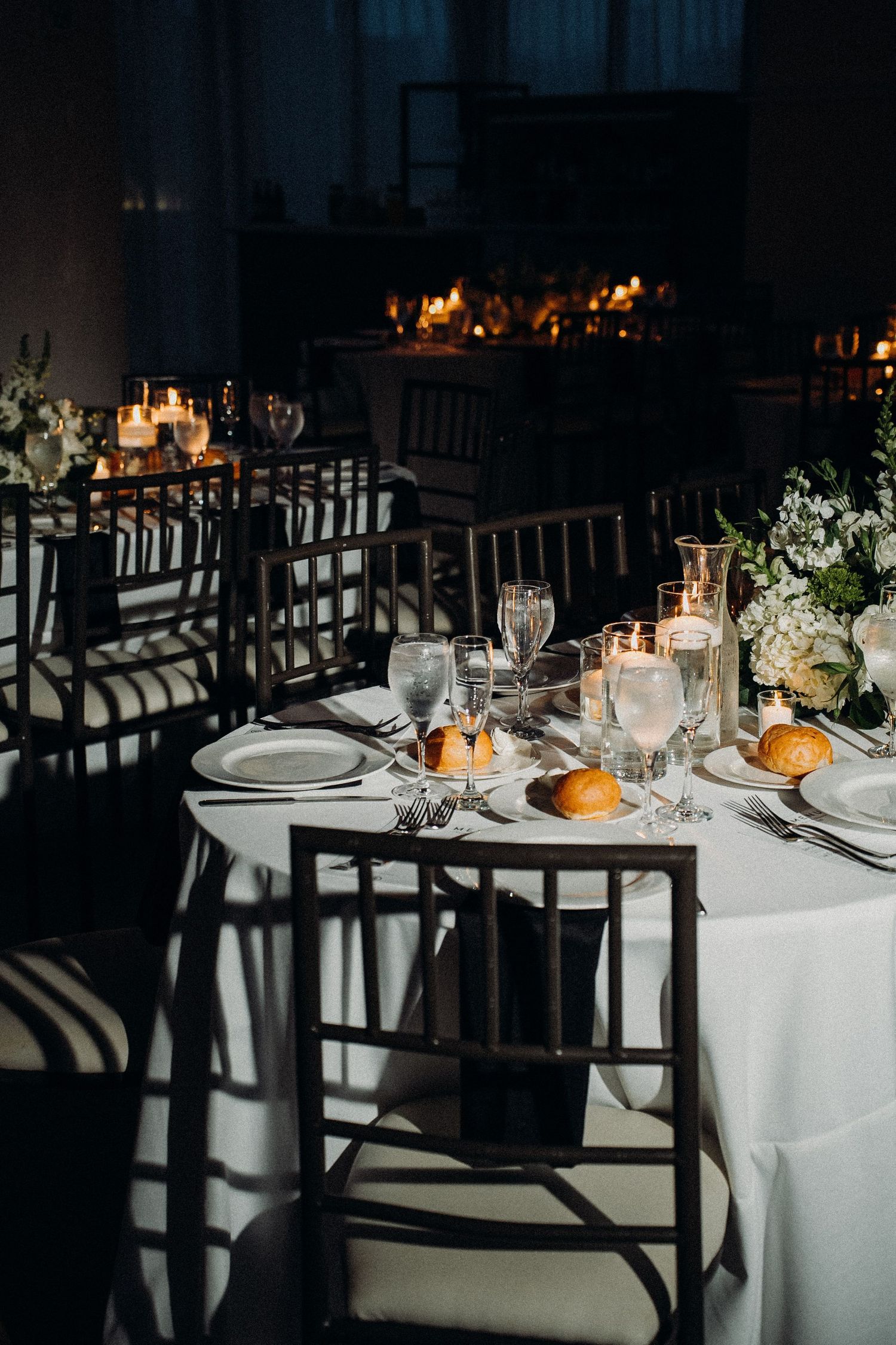 Elegant wedding reception tables set with white linens, glassware, and dark wooden chairs casting dramatic shadows.