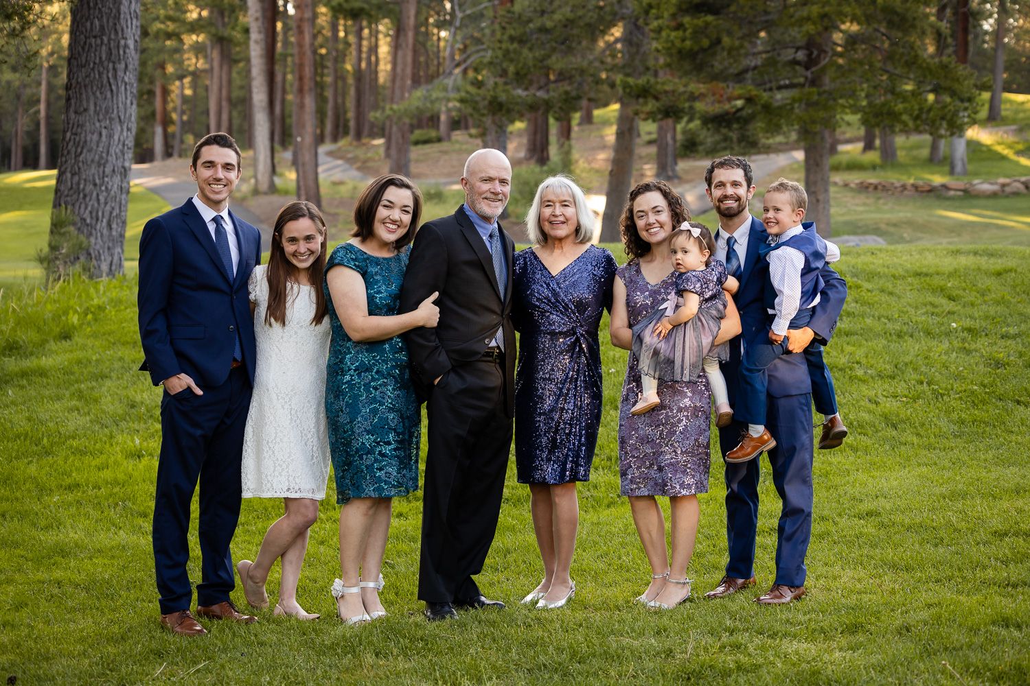 A formally dressed extended family session with people standing together outdoors in a wooded area during golden hour.