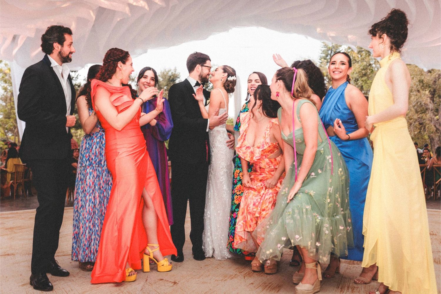 A group of wedding guests in colorful formal attire gather and laugh together during an outdoor celebration.
