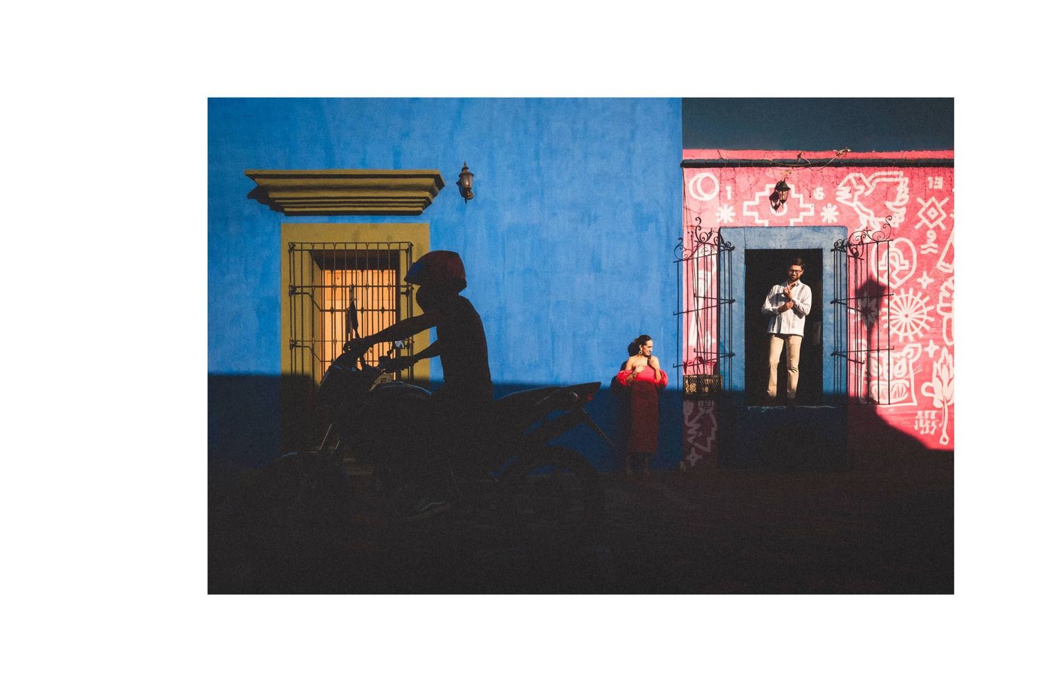 Colorful building facades in blue and pink with silhouetted figures standing in doorways casting long shadows.