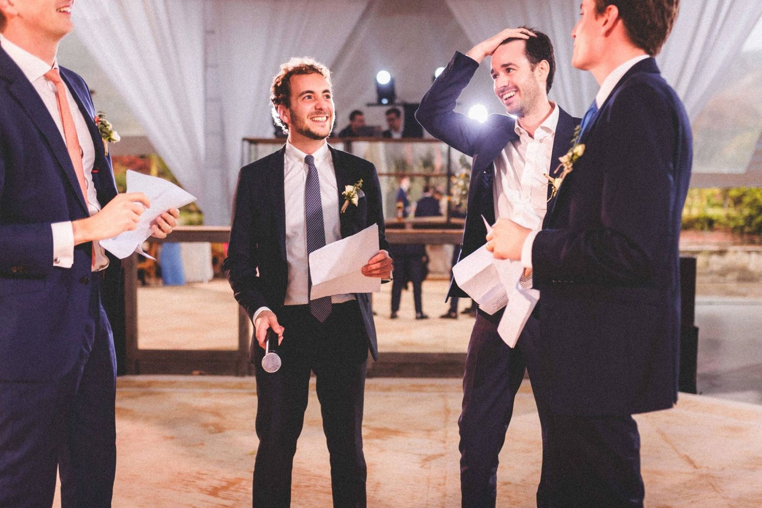 Groomsmen in black suits share a lighthearted moment while reading papers at an elegant outdoor wedding venue.