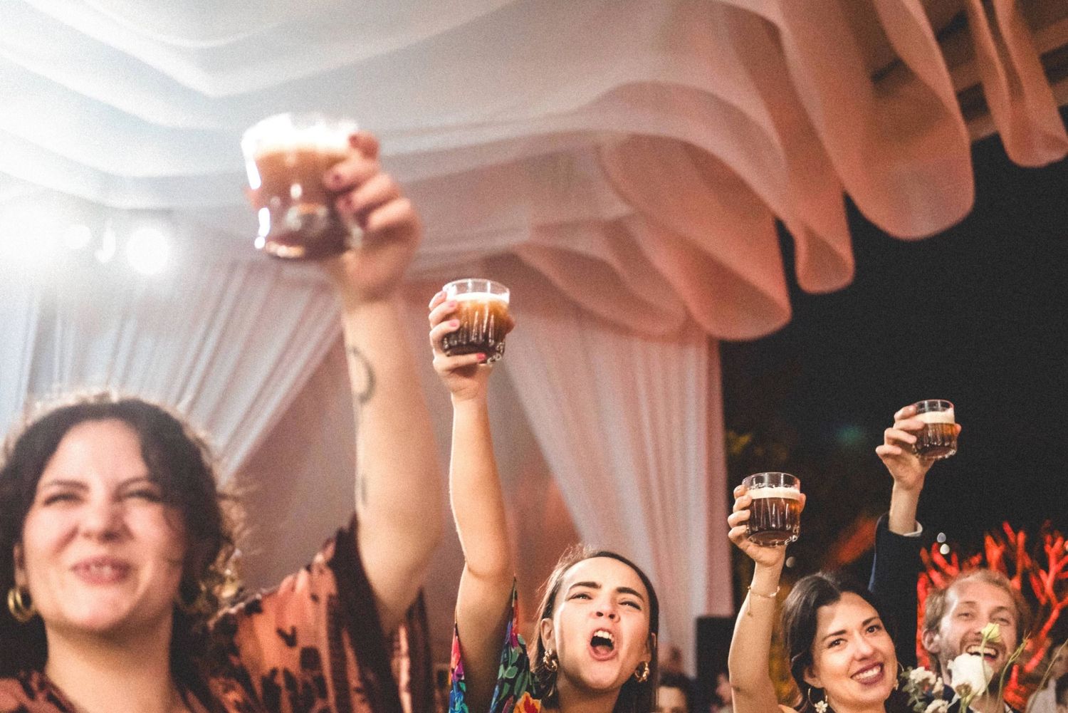 Enthusiastic wedding guests raise their fists in celebration under flowing white tent draping.