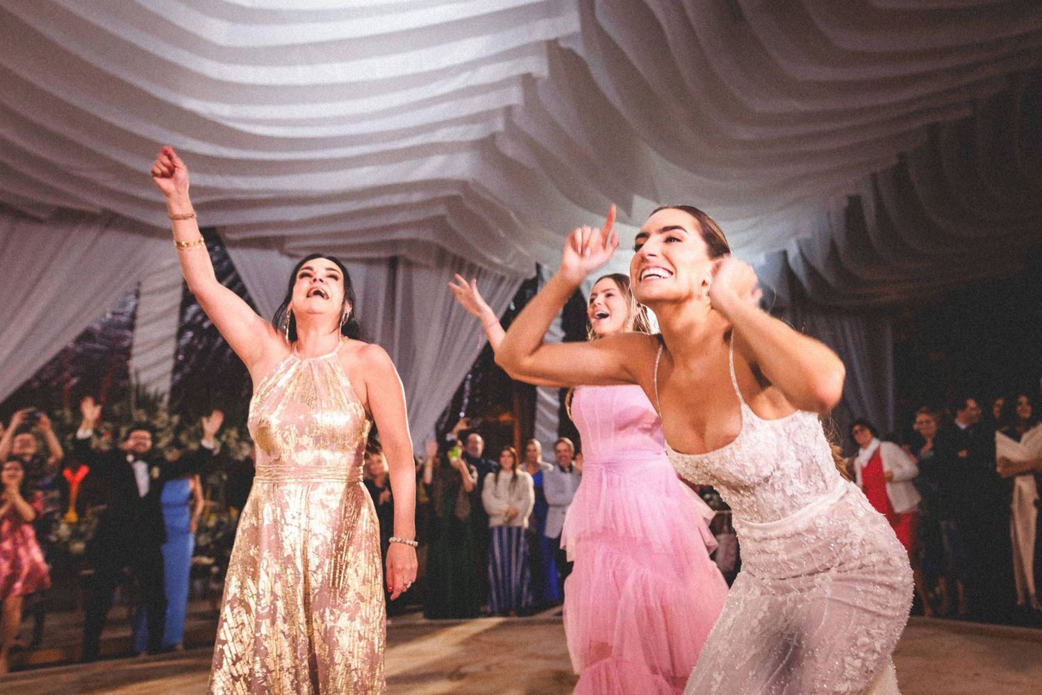 Wedding party dancing joyfully under white draped ceiling at reception.