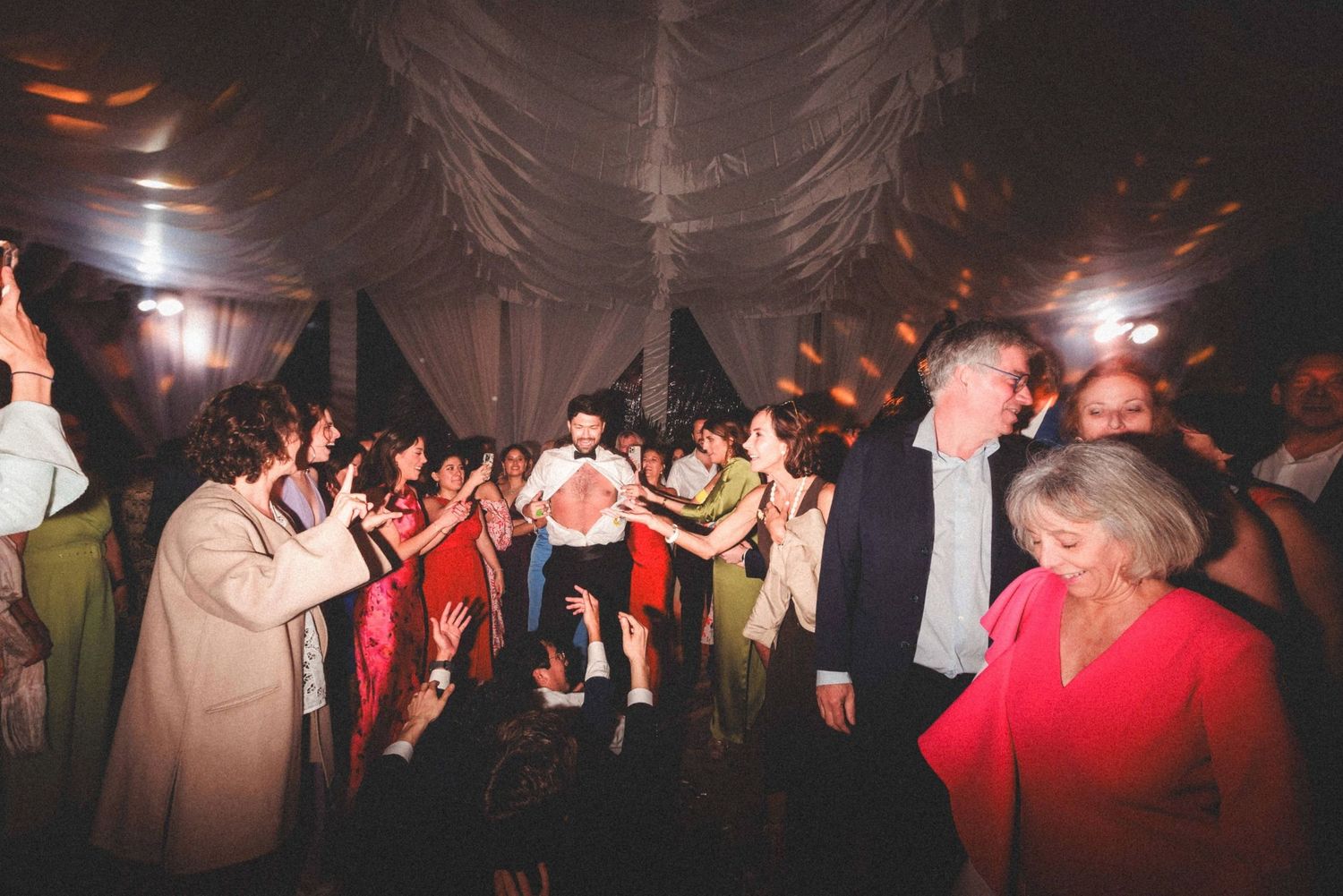 A crowd of people dancing and celebrating at a dimly lit party with atmospheric lighting.