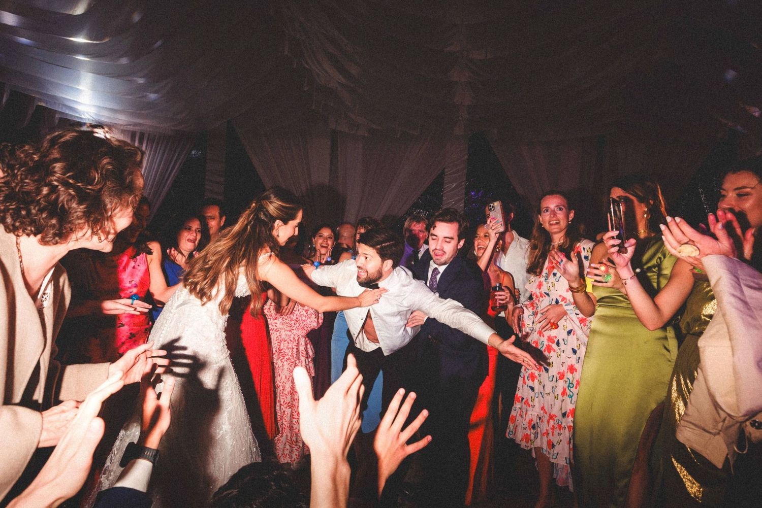 A group of wedding guests dance and celebrate on a dimly lit dance floor at a reception party.