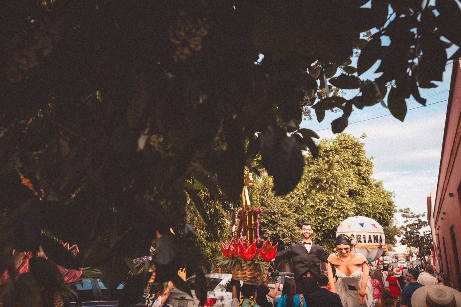 Colorful festival parade with red banners and flags moving through a tree-lined street.