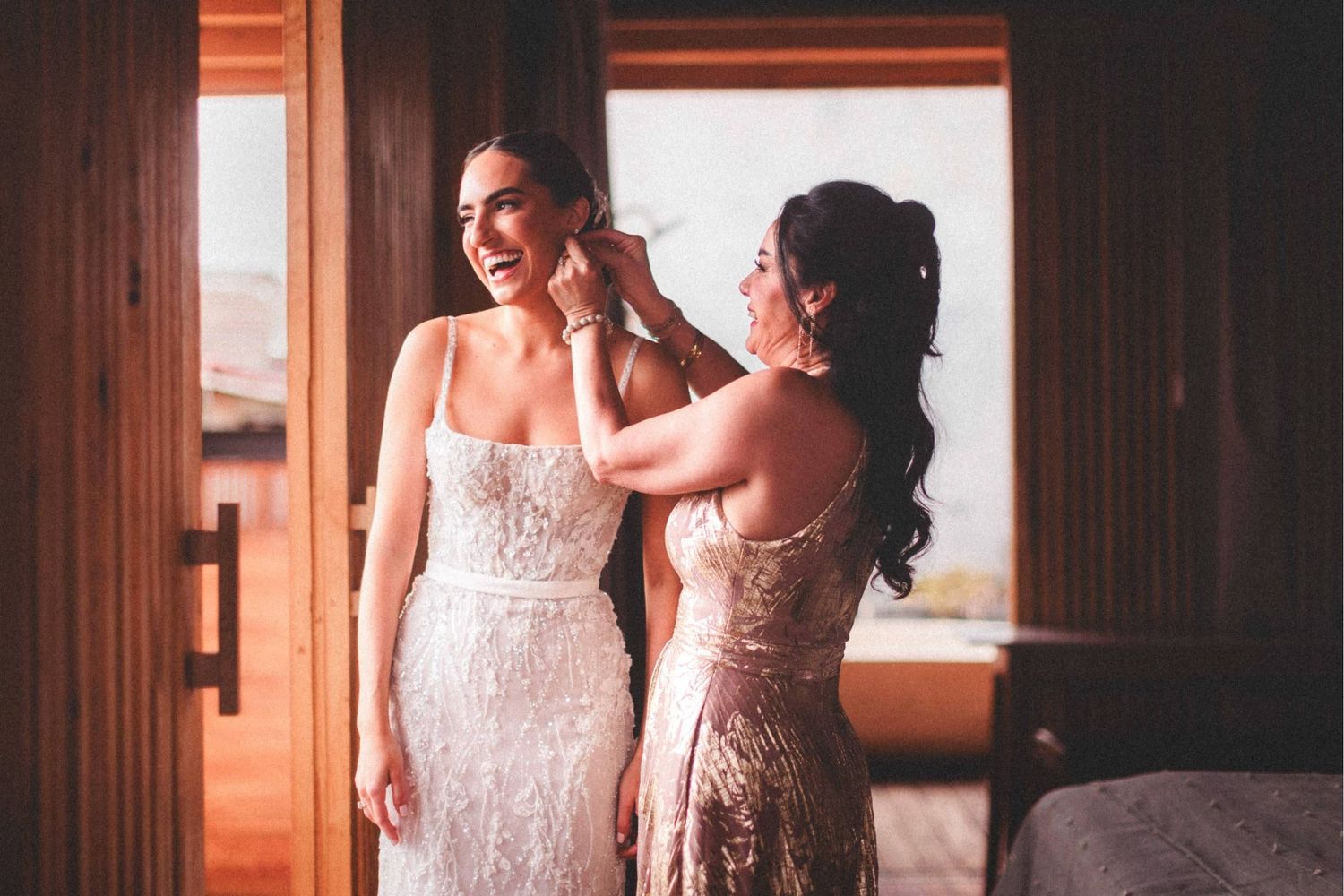 Two people in elegant dresses share an intimate moment in a warmly lit interior setting.
