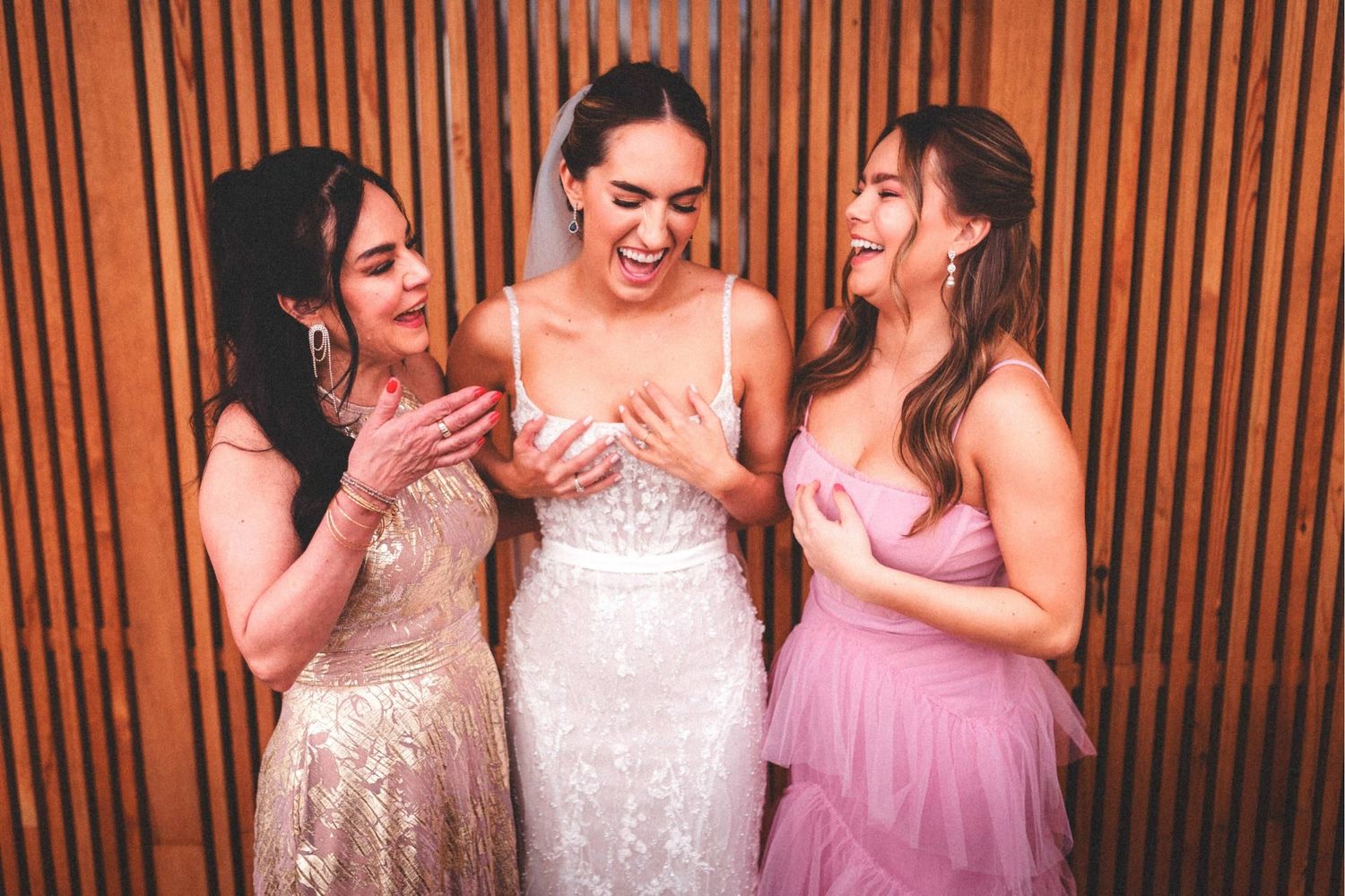 Three friends in formal dresses share a joyful moment laughing together against a wooden panel background.