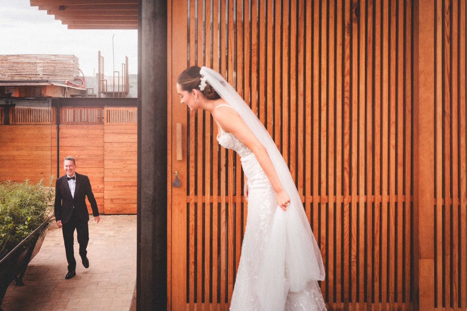 Wedding couple walking along modern wooden slatted wall in outdoor venue setting.