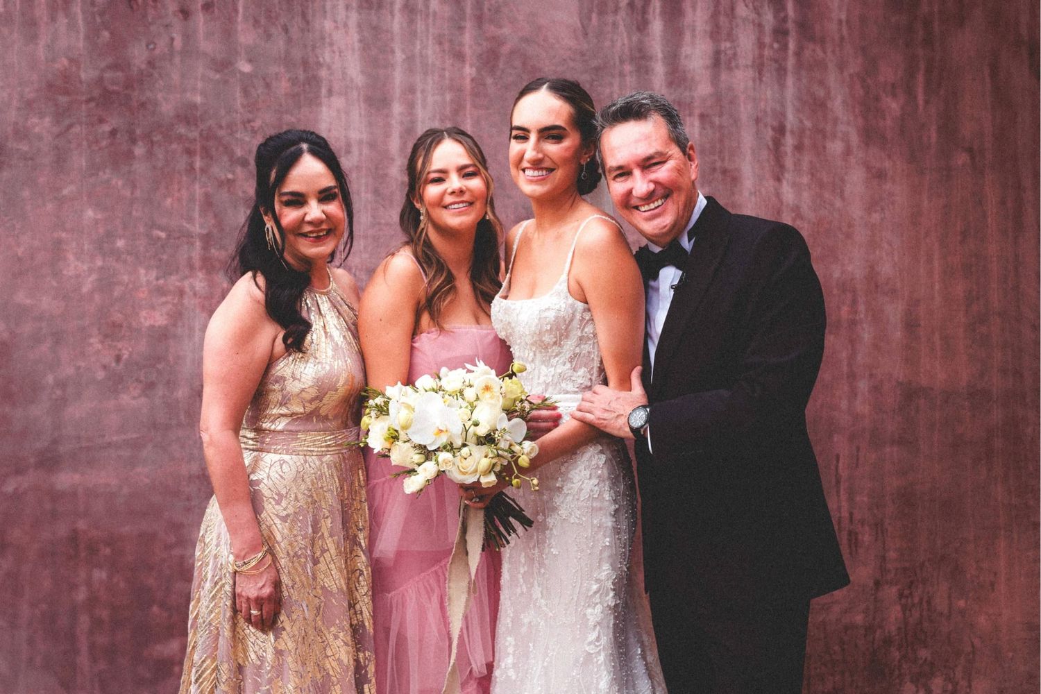 Wedding party in formal attire poses together with bouquet against textured burgundy wall.
