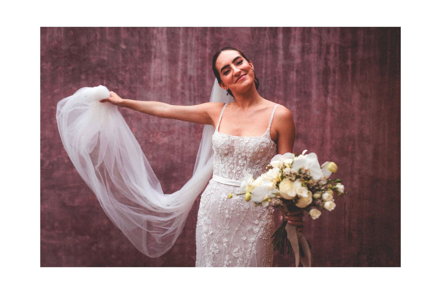 A bride in a beaded wedding gown poses with a flowing veil and white floral bouquet against a burgundy backdrop.