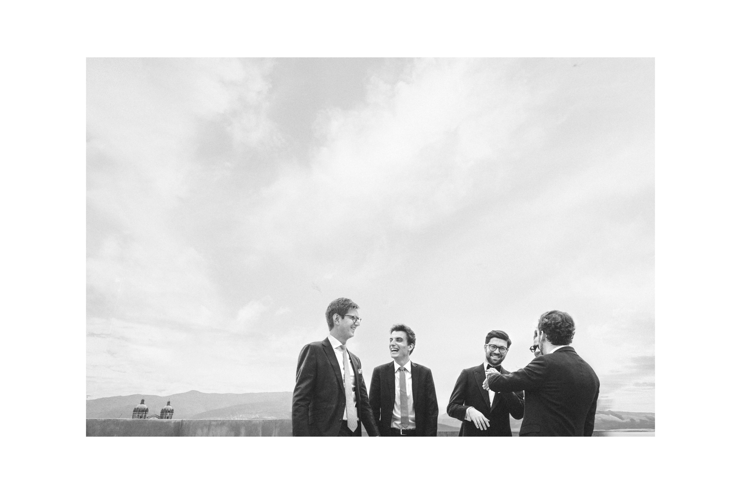 A black and white photo shows a group of formally dressed people standing together outdoors against a cloudy sky.