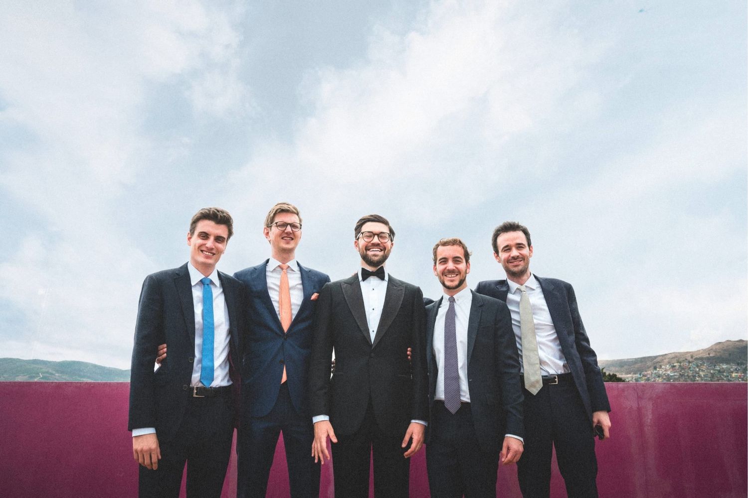 Wedding party members standing outdoors against cloudy sky in formal attire.